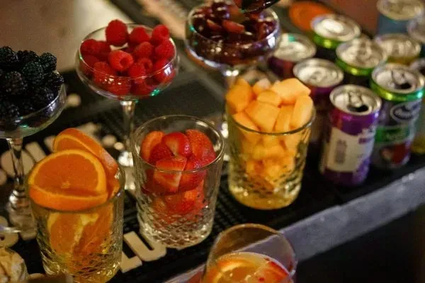 Colorful fresh fruit garnishes in glasses with blurred drink cans.