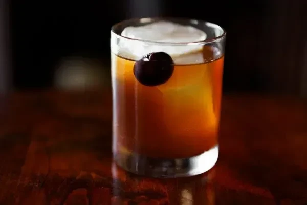 Dark cocktail in a rocks glass with large ice and a cherry.