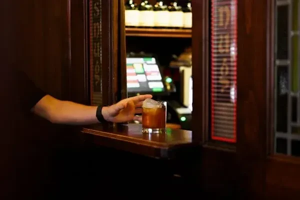 Hand reaches for amber iced drink at a dark wooden bar.