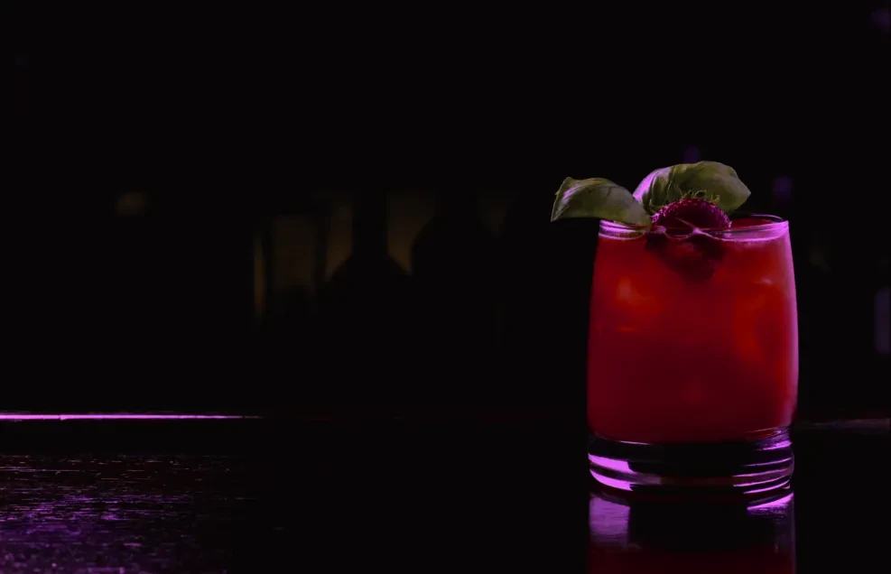Reddish-pink cocktail with strawberry and basil in a dramatic, dark bar.