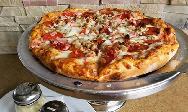 Large meat-topped pizza on a stand with shakers and stone wall.