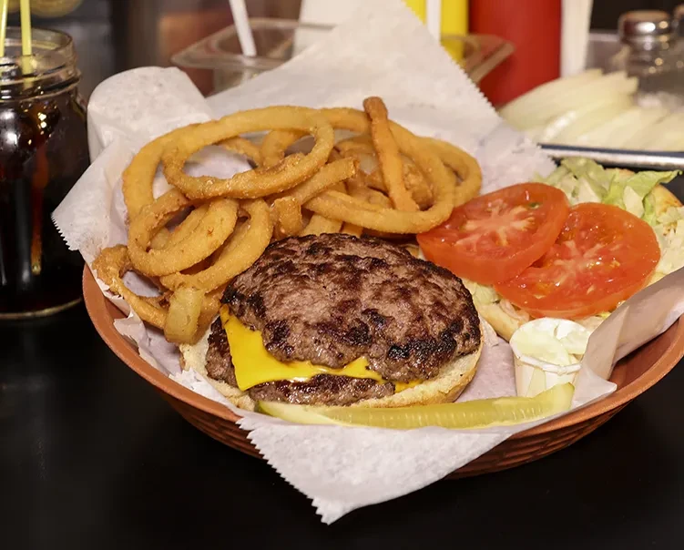 Cheeseburger with melted cheese, onion rings, pickle, and soda in basket.
