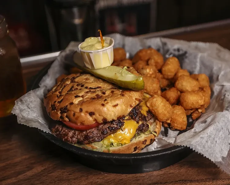 Burger topped with a pickle spear, tater tots, and dipping sauce.