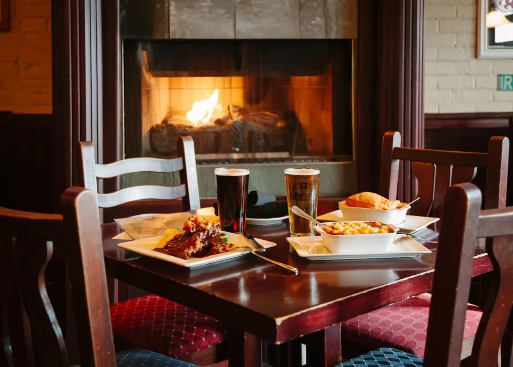 Cozy restaurant table with hearty food, beers, and a roaring fireplace.