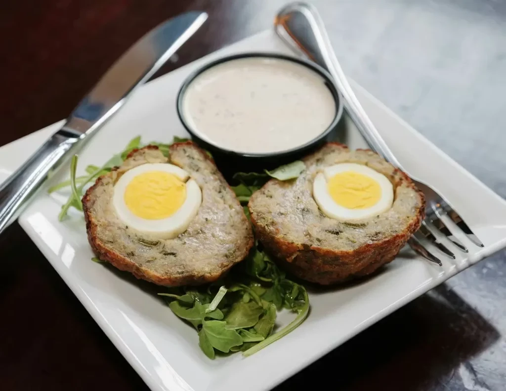 Two halved, golden Scotch eggs with visible yolks, dipping sauce, and greens.