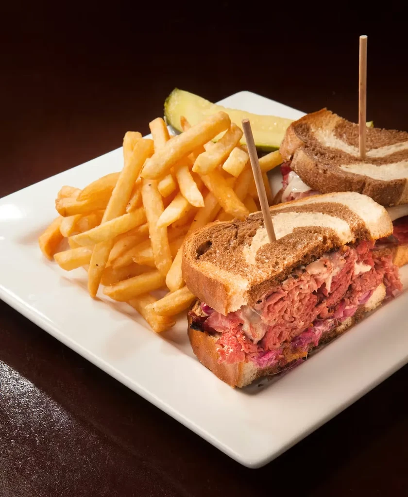 Reuben sandwich, french fries, and pickle on a white plate.