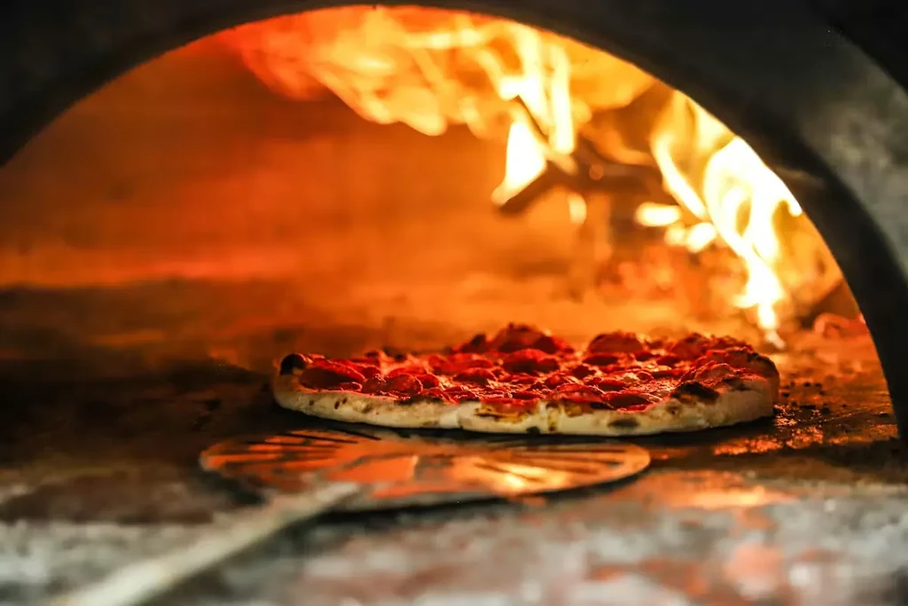 Pepperoni pizza baking in a wood-fired oven with visible flames.