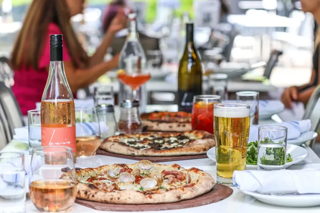Three pizzas, rosé wine, beer, and drinks on a lively restaurant table.