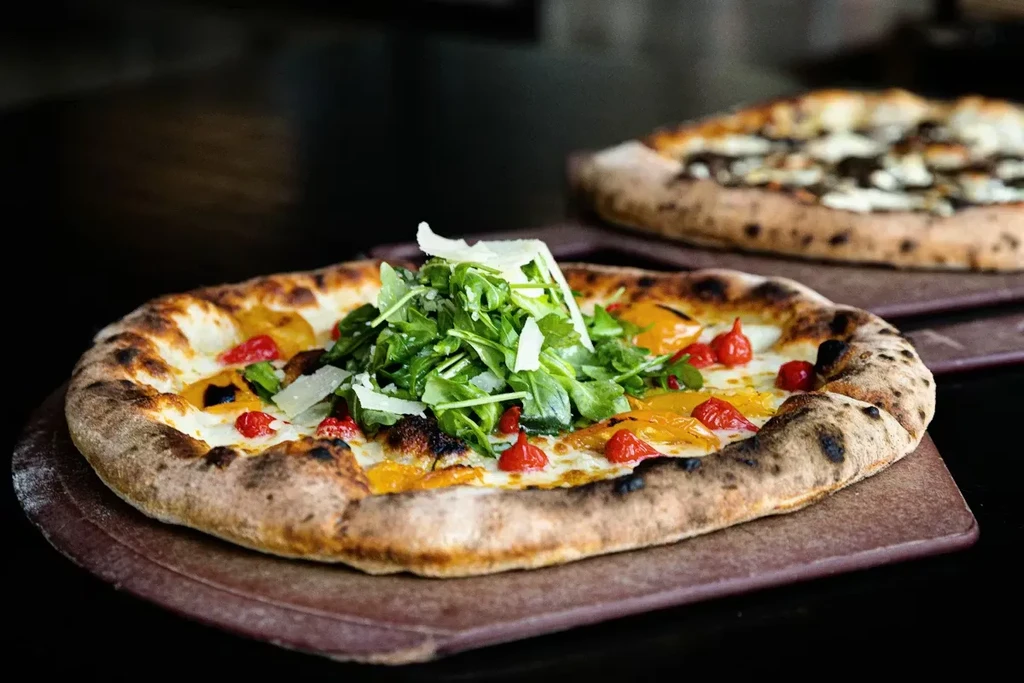Wood-fired pizza with arugula, parmesan, red peppers; another blurred in back.