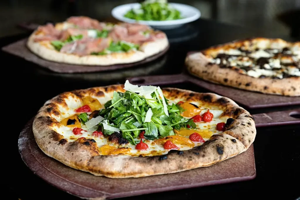 Three Neapolitan pizzas on wooden peels; one with peppers, arugula, cheese.