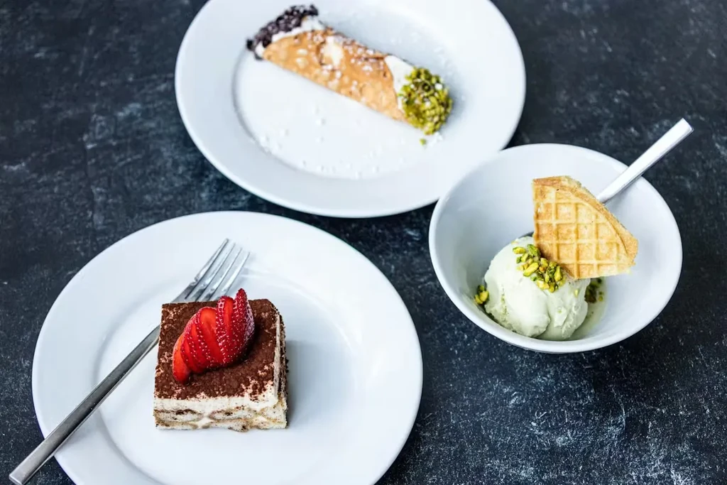 Cannoli, tiramisu with strawberry, and pistachio ice cream on dark surface.