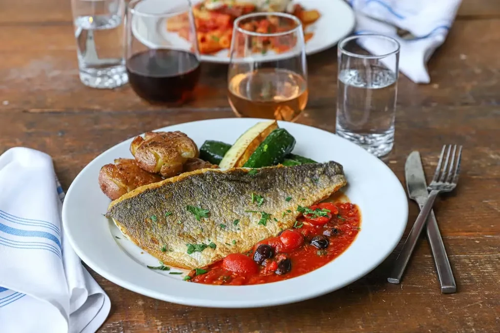 Whole pan-seared fish, tomato-olive sauce, potatoes, zucchini on wooden table.