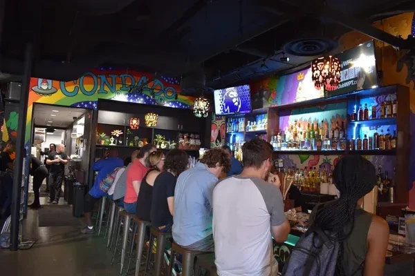 Busy bar with illuminated liquor, colorful murals, and "CONDADO" sign.
