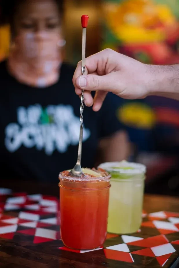 Hand stirs a garnished reddish-orange drink in a mason jar.