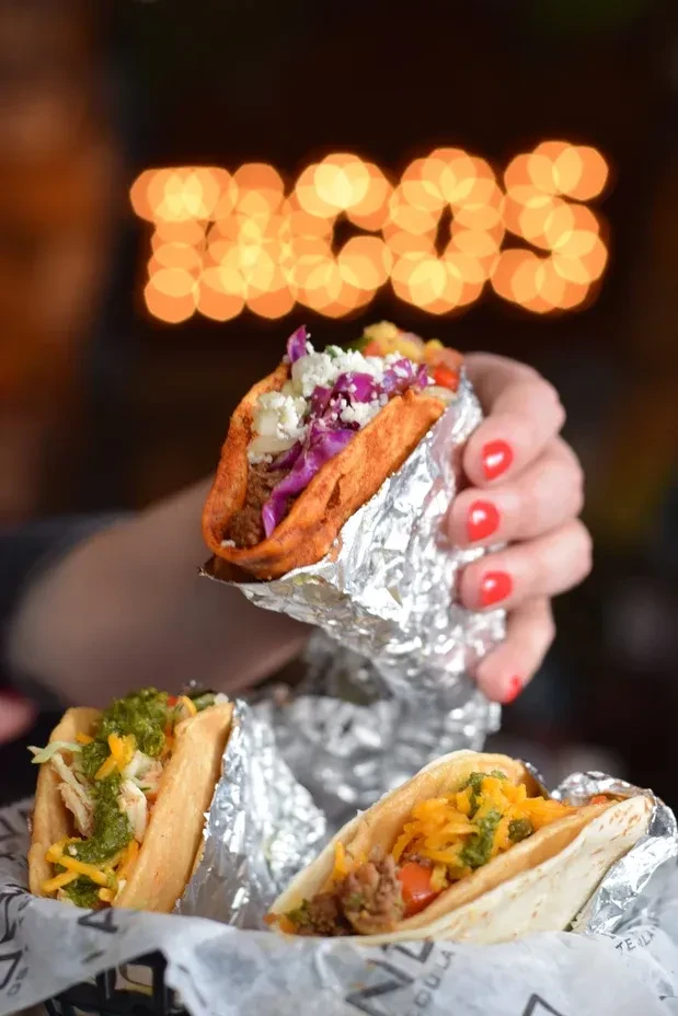 Three tacos: one held by hand, two in basket. TACOS sign blurred.