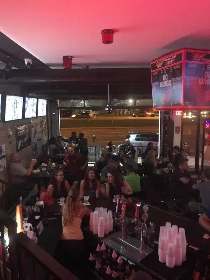 Red-lit, lively bar interior with patrons, bartender, and sports screens.