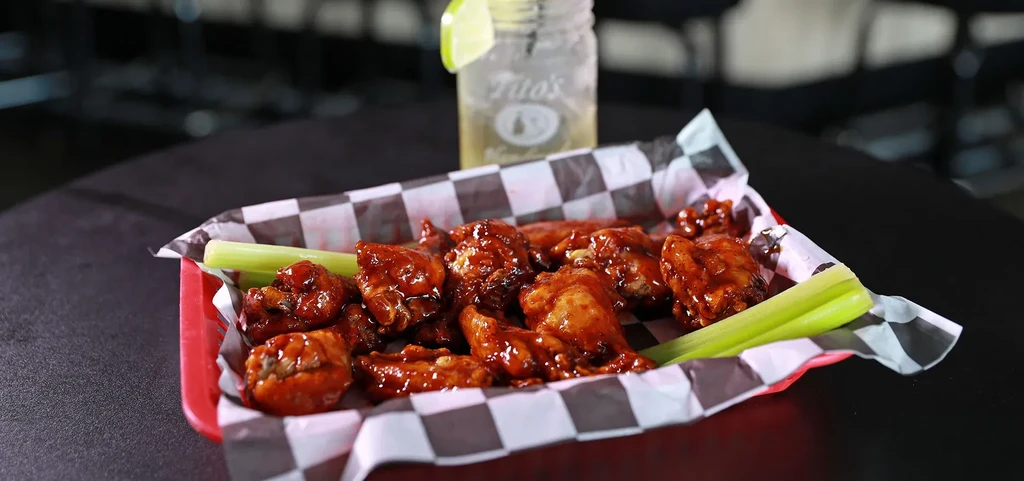 A red basket of saucy chicken wings, celery, and a Tito's drink.