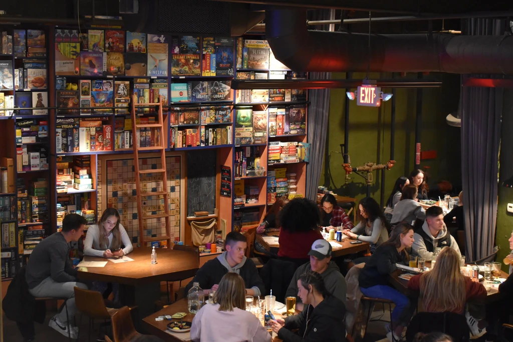 Bustling board game cafe with industrial decor, shelves of games, and patrons.