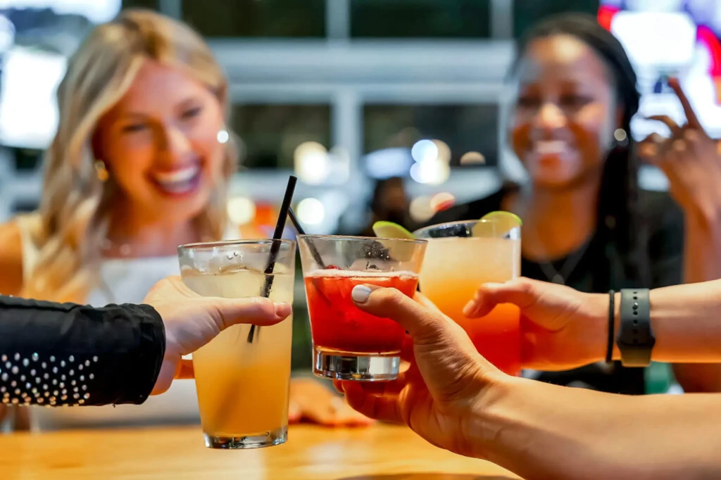 Three people toast colorful drinks; two smiling women in background.