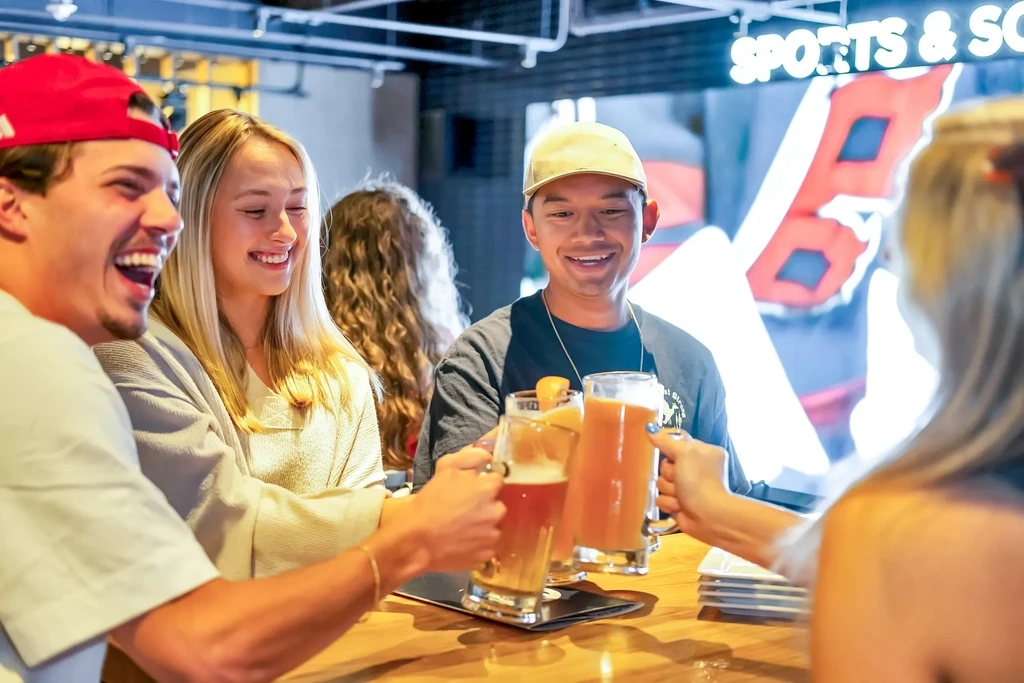 Four young adults laughingly toast drinks at a casual sports bar.