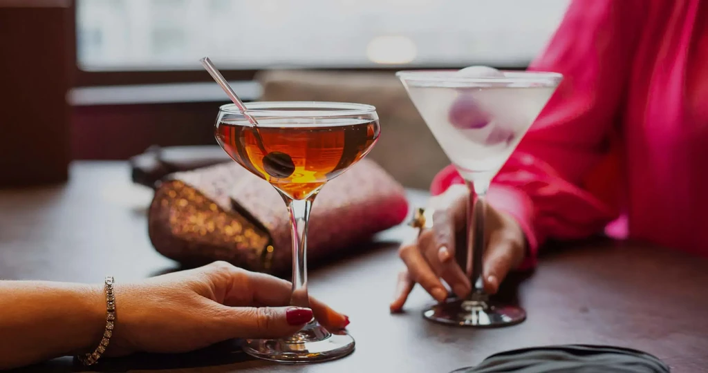 Two hands hold cocktails in coupe and martini glasses on a dark table.