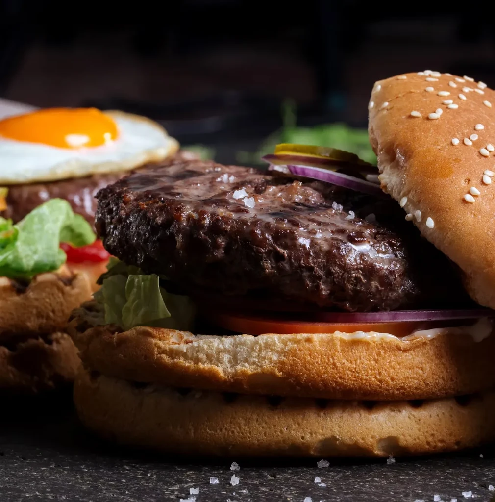 Close-up of a beef burger with lettuce, tomato, onion; egg burger behind.