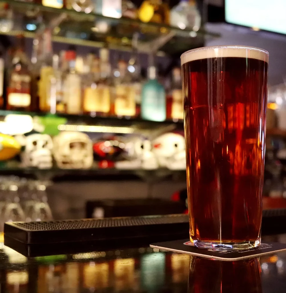 Full glass of amber beer on a bar, with liquor bottles and helmets.