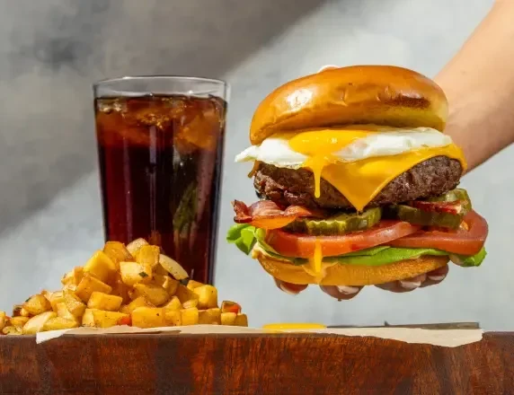 Hand holding a stacked burger with egg, cola, and roasted potatoes.