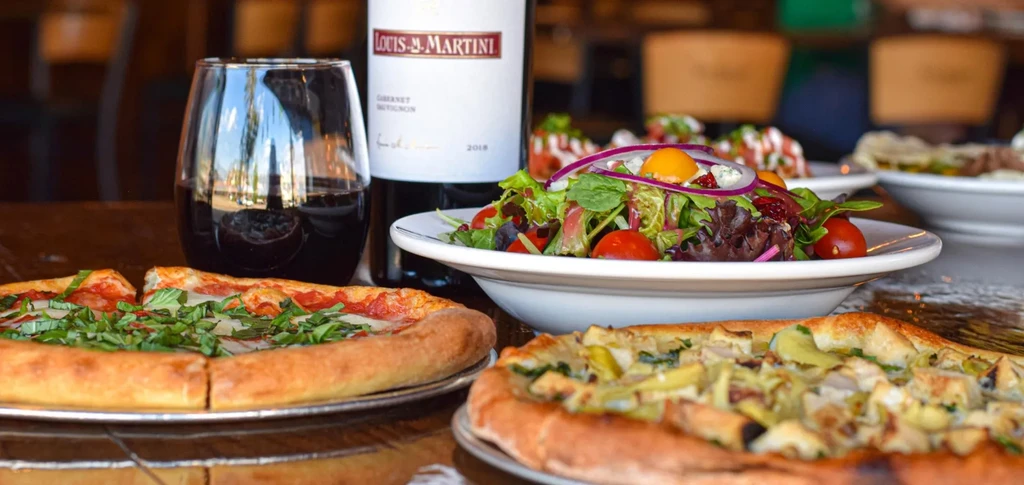 Restaurant table with two pizzas, Louis M. Martini Cabernet, and salad.