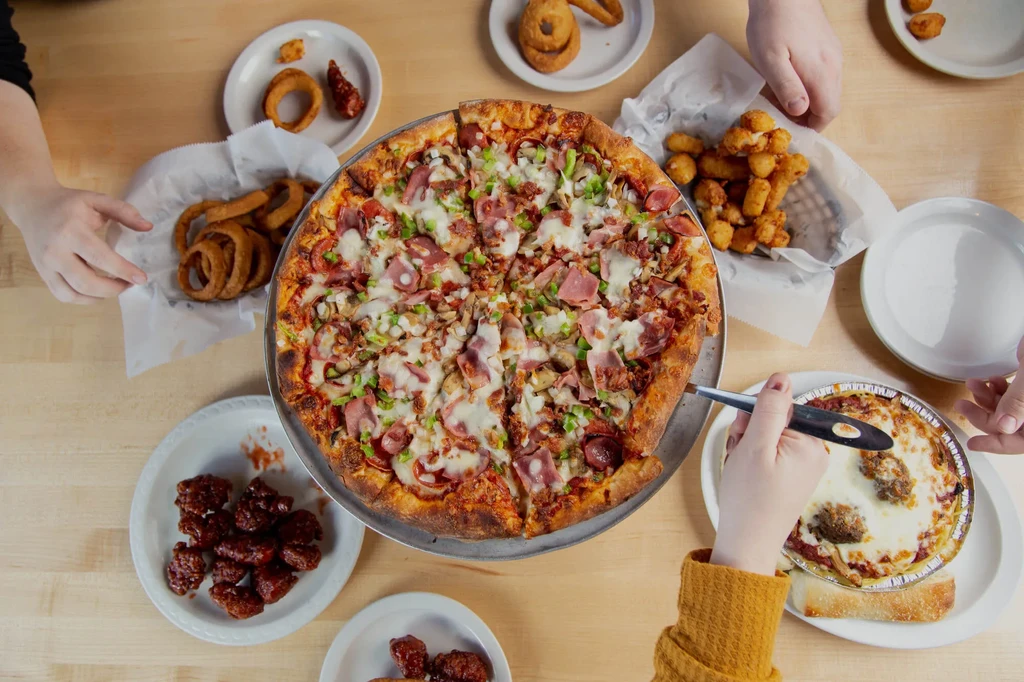 Table with hands sharing pizza, wings, pasta, and fried sides.
