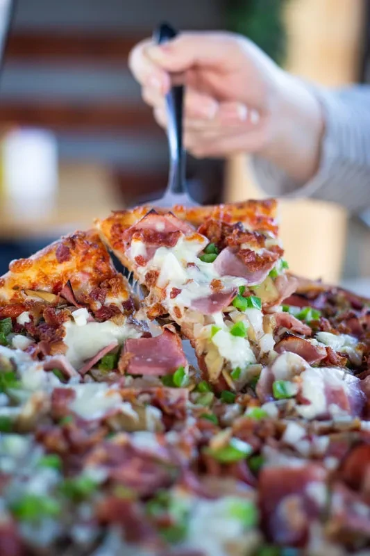 Hand lifting a slice of ham, bacon, and bell pepper pizza.