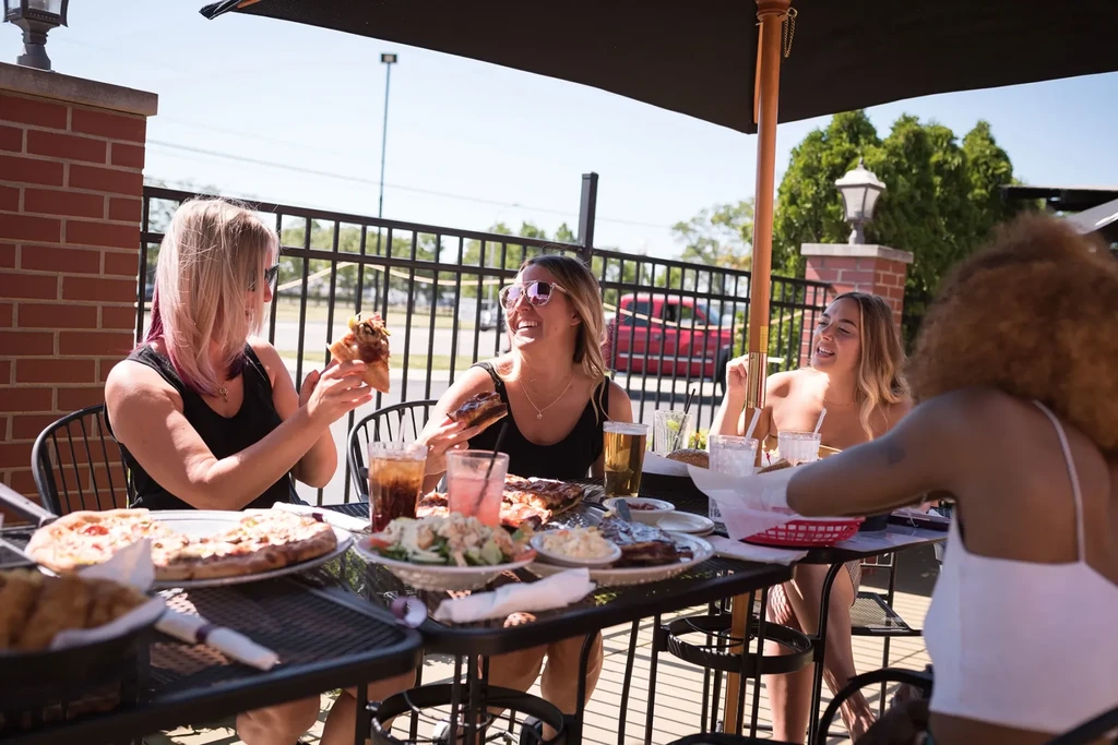 Four women enjoy a diverse meal at a sunny outdoor patio.