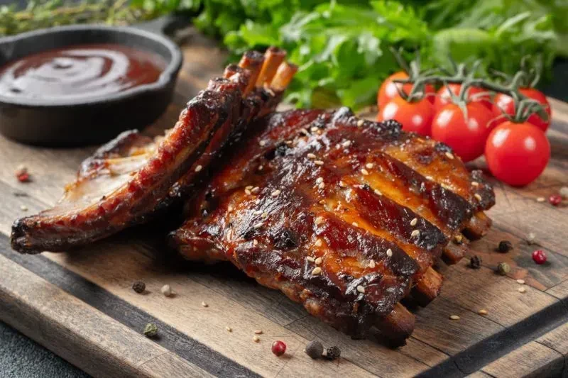 Glossy barbecue ribs on a wooden board with sauce, tomatoes, and herbs.
