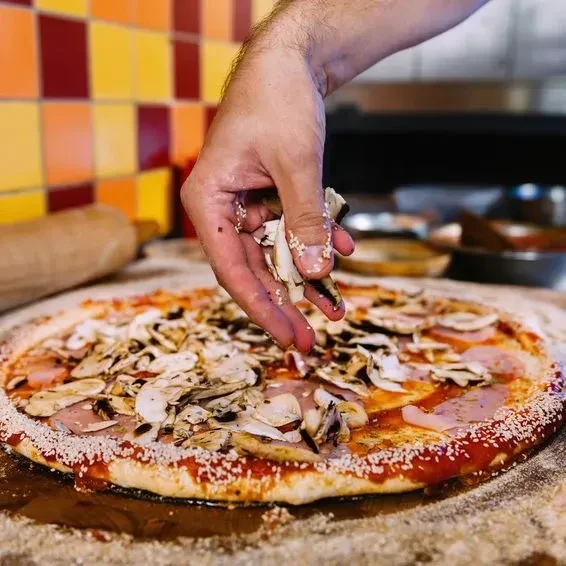 Hand sprinkling toppings on uncooked pizza with meat, mushrooms, sesame crust.