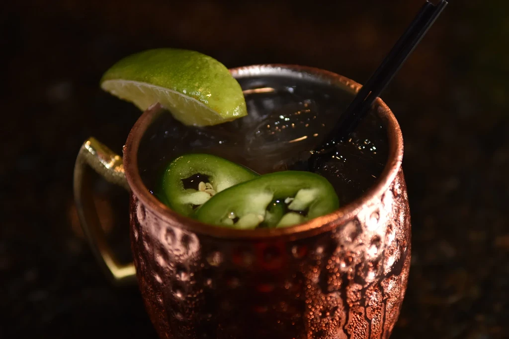 Dark drink in hammered copper mug with ice, lime, and jalapeño slices.