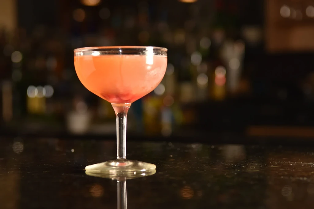 Reddish-pink cocktail with small pieces in coupe glass on bar.