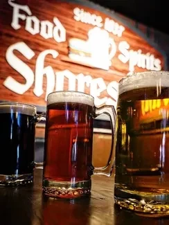 Three distinct beers in mugs, a blurred "Food Spirits Shores 1887" sign.