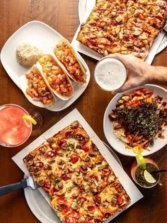 Overhead shot: pizzas, tacos, grain bowl, cocktails, and beer on table.