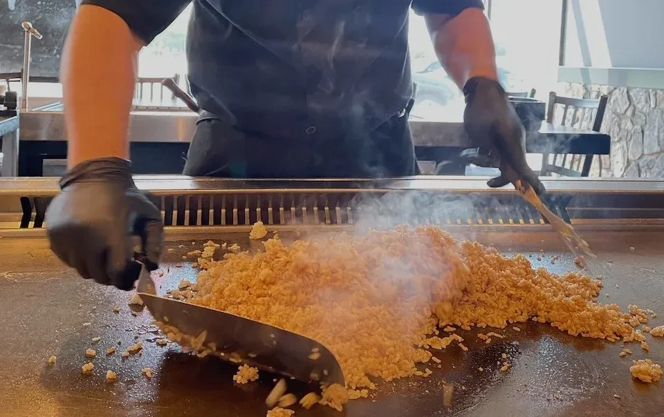 Chef's gloved hands prepare steaming fried rice on hibachi grill.