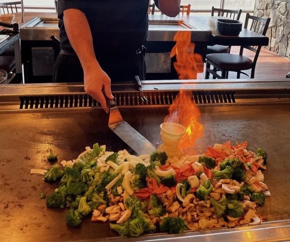 Chef cooking colorful vegetables on teppanyaki grill with dramatic flames.