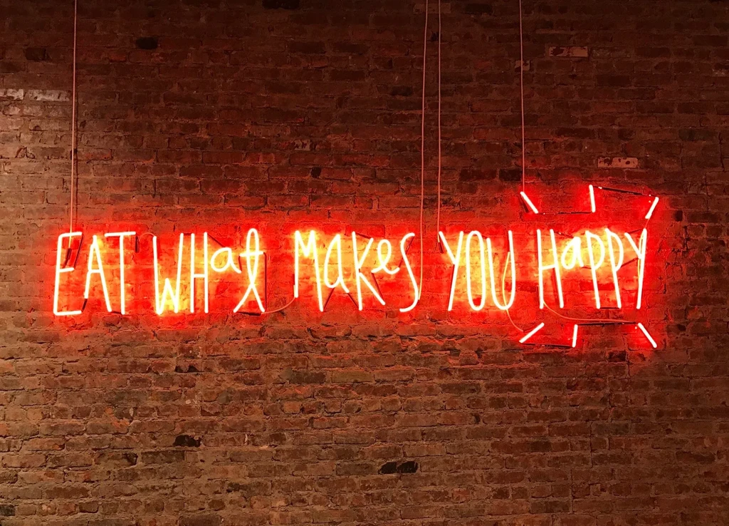 Red neon sign "EAT WHAT MAKES YOU HAPPY" against a brick wall.