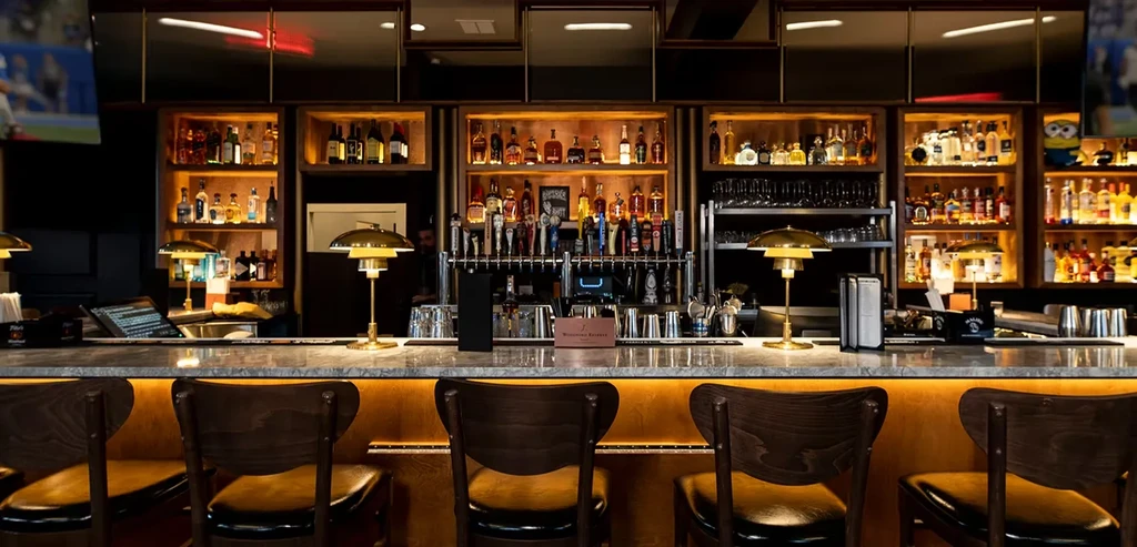 Modern bar with marble counter, dark stools, and extensive illuminated liquor display.