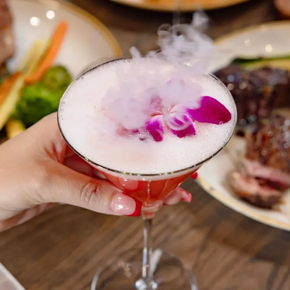Pink cocktail with foam, purple orchid, and smoky mist in martini glass.