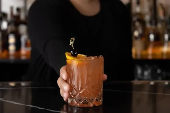 Hand holds rustic-orange cocktail in cut-glass with ice, orange slice, cherry.