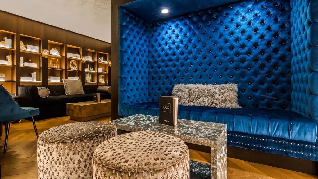 Elegant lounge with blue velvet seating, patterned ottomans, and illuminated shelving.