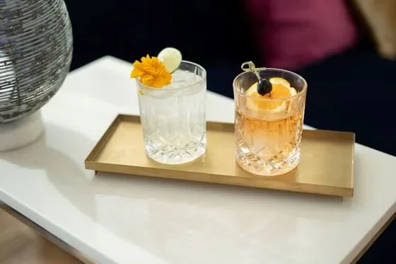 Two distinct garnished cocktails in ornate glasses on a gold tray.