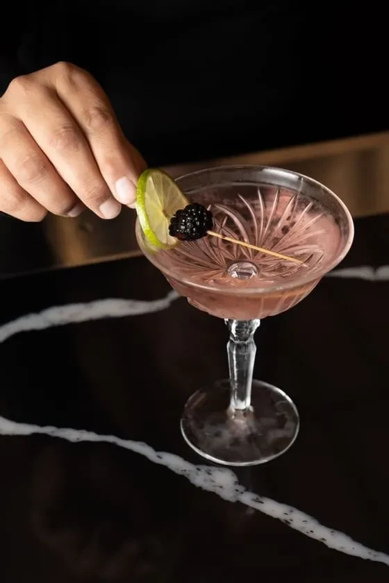 Hand adjusts reddish-pink cocktail with lime and blackberry garnish on marble.