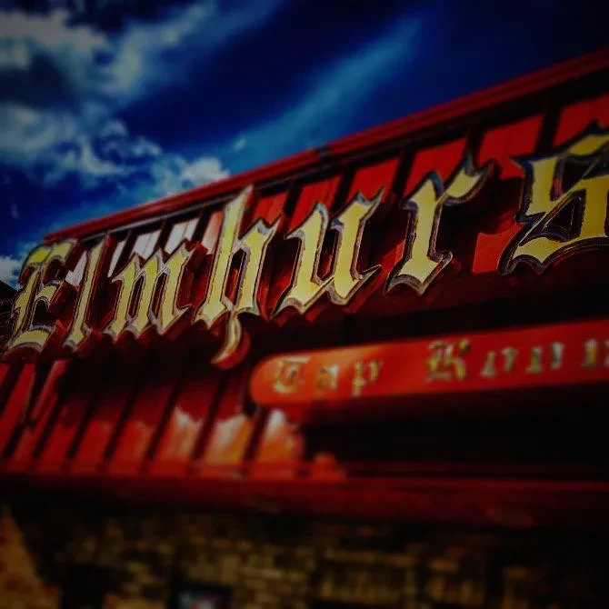 Golden "Elmhurst" sign on red ribbed building, under a cloudy blue sky.