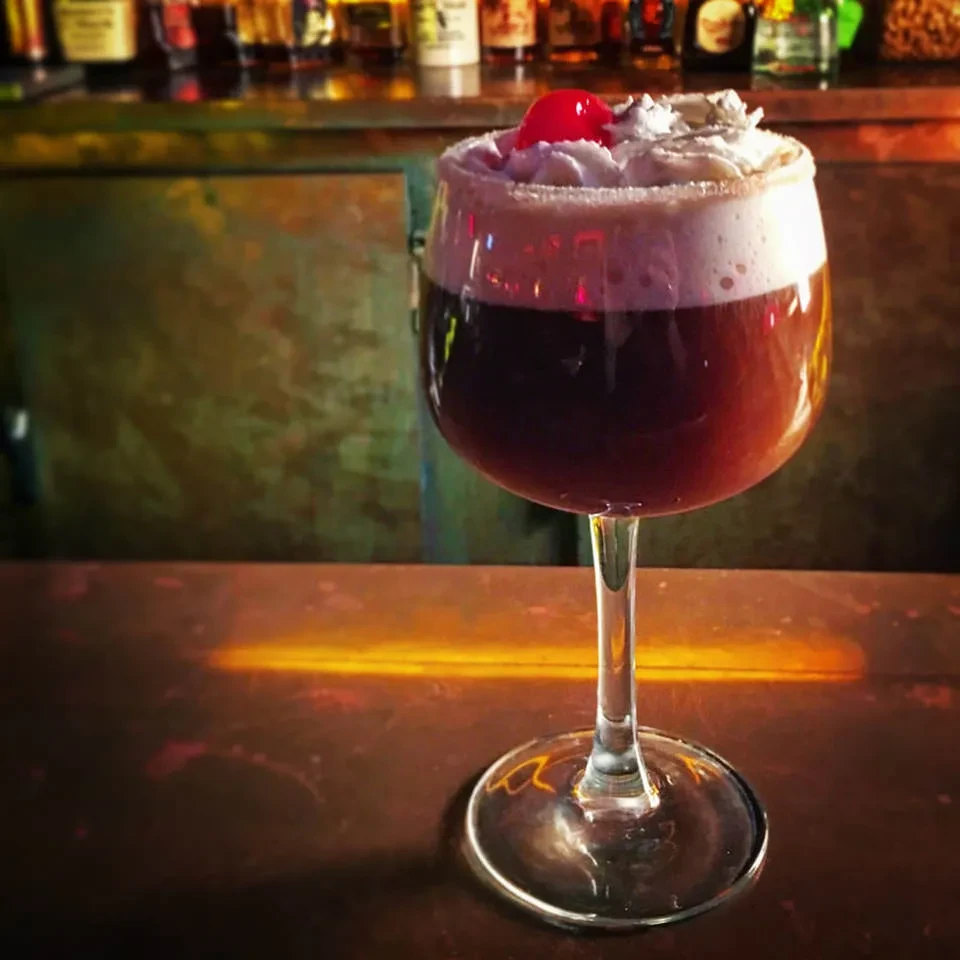 Dark red cocktail with whipped cream, cherry, and sugared rim on bar.