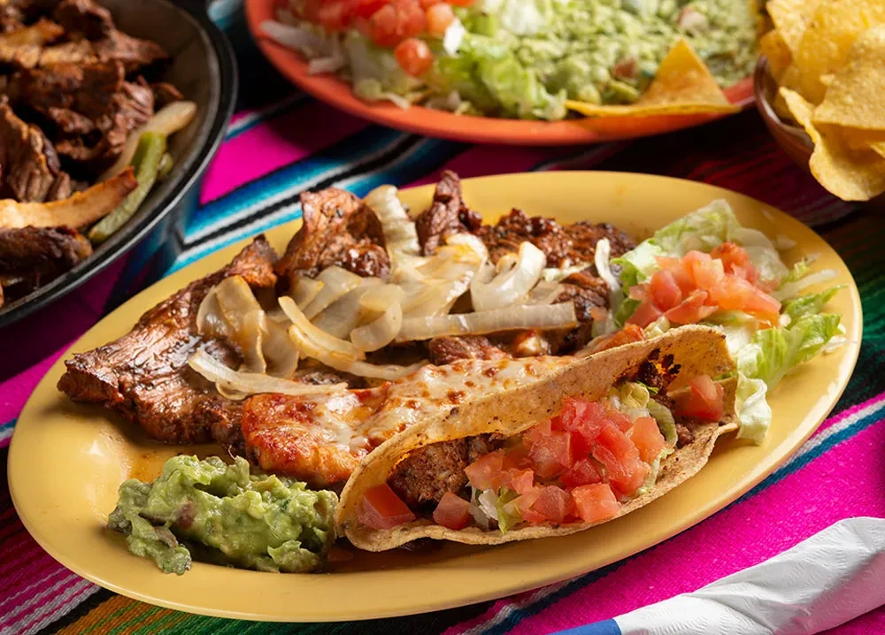 Grilled meat with cheese, taco, guacamole on a vibrant Mexican blanket.